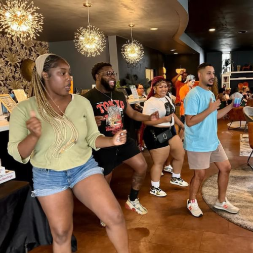 dancing-group A group of individuals sitting around a table in a meeting or conference setting. Behind them is a poster board with various texts and logos, suggesting an event like a workshop or presentation. Laptops and papers are on the table, indicating collaborative work or discussion.