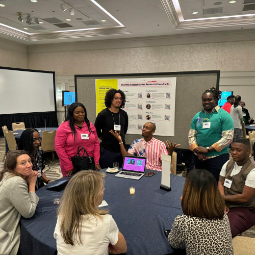 group-desk A group of individuals sitting around a table in a conference or meeting setting. Behind them is a poster board with various texts and logos, suggesting an event like a workshop or presentation. Laptops and papers are on the table, indicating collaborative work or discussion.