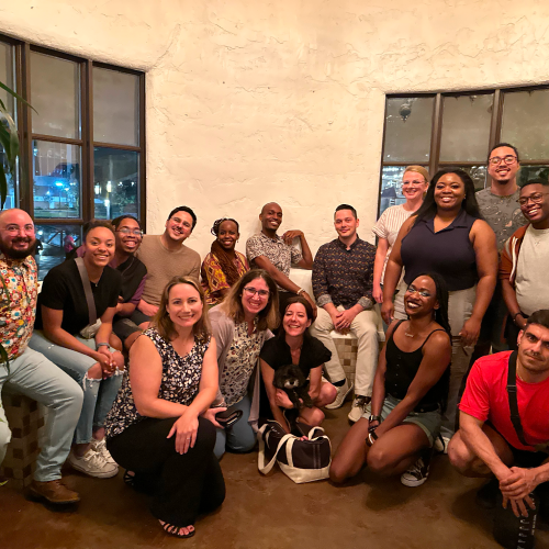 group-photo A group of individuals sitting and standing together in a room with white walls and windows.