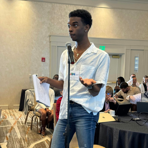 guy-asking An individual standing in front of a microphone, holding papers in one hand and gesturing with the other.