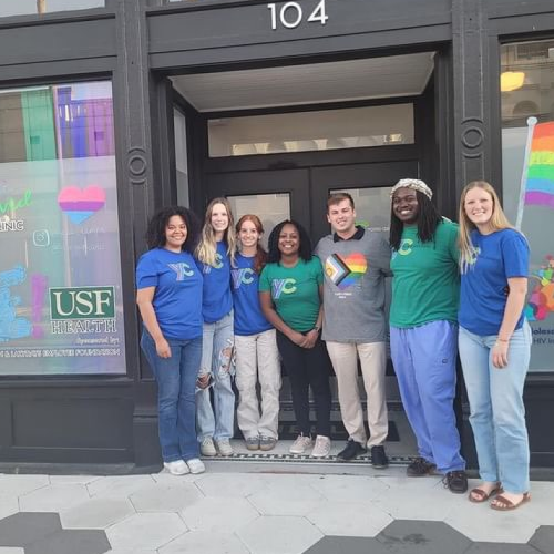 outside-group A group of seven people standing in front of a building entrance marked with the number 104. They are wearing shirts with various designs, including rainbow motifs and logos, possibly indicating support for the LGBTQ+ community or a related event.